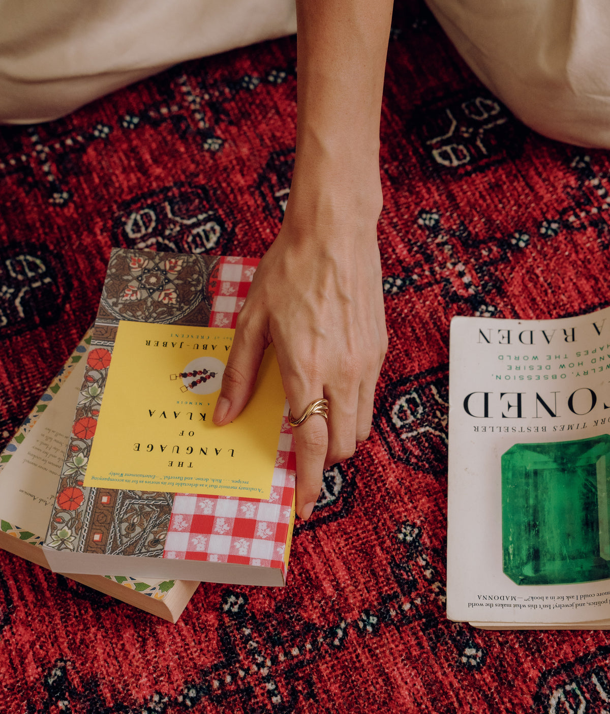 A hand wearing a gold ring resting on a stack of books on a red rug